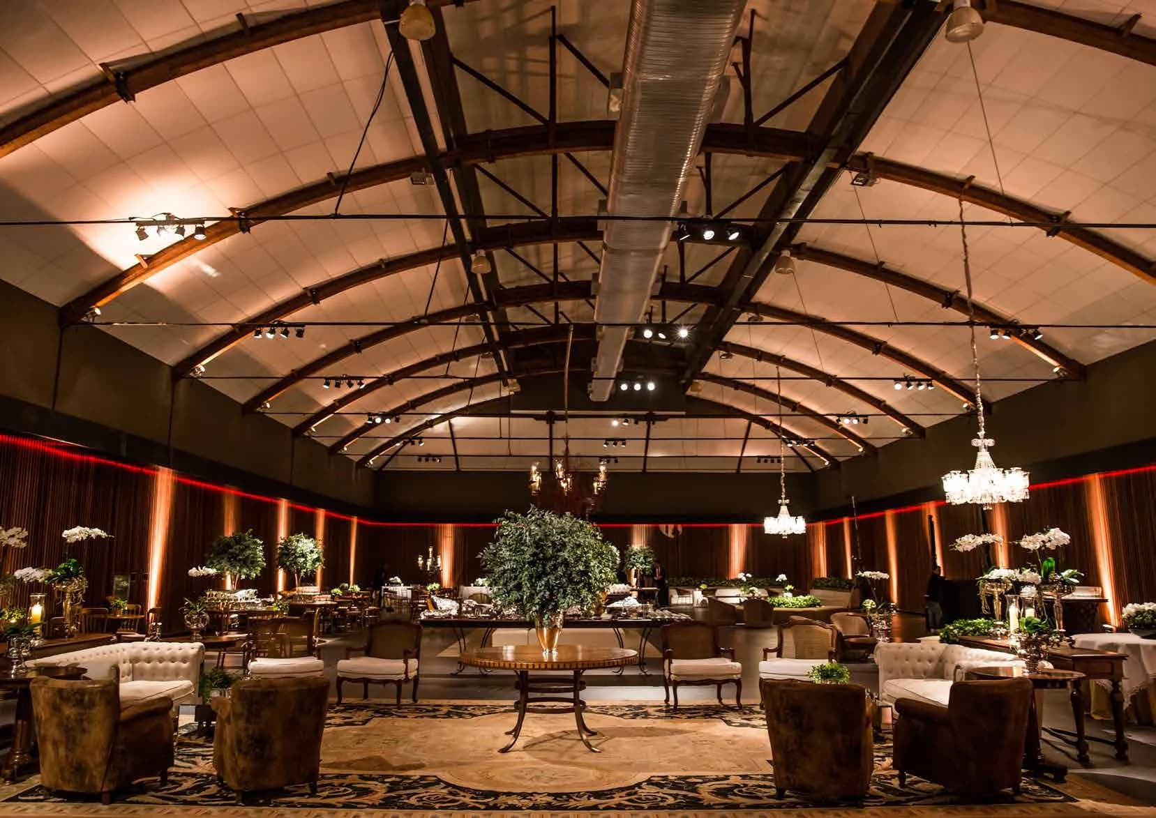 Espaço de recepção com lustres, lounges e decoração floral sob um teto de estrutura aparente
